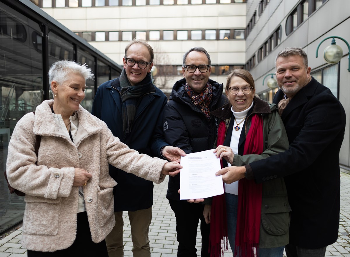 Fem personer står utanför en byggnad. Alla håller i ett papper. De är glada.