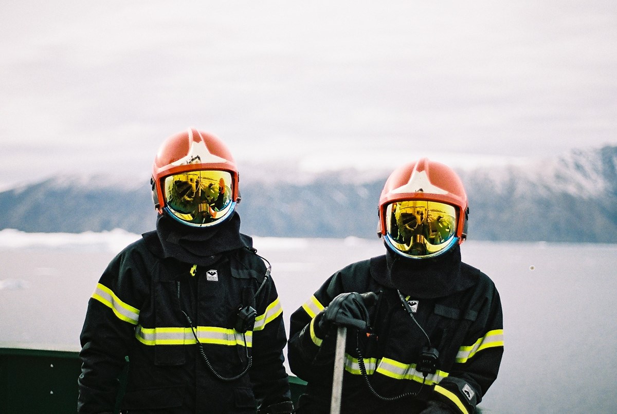 Två personer iklädda brandutrustning och hjälmar med visir, tittar in i kameran. I bakgrunden ett berglandskap