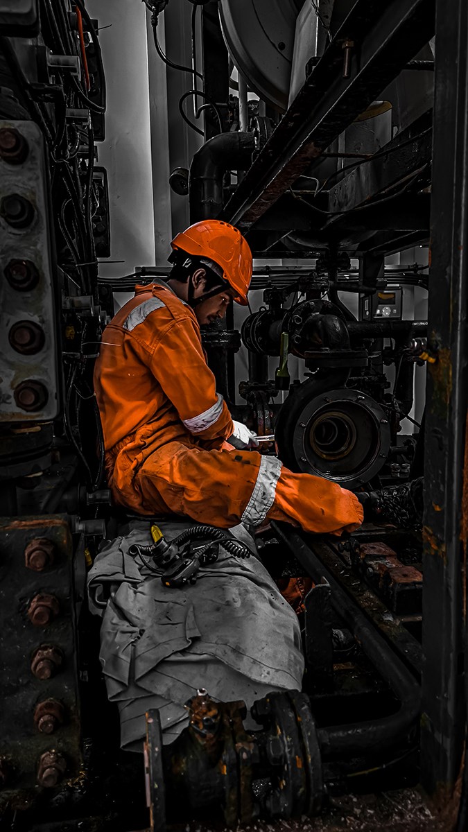 A worker dressed in an orange coverall and helmet sits in the darkness of an engine room.