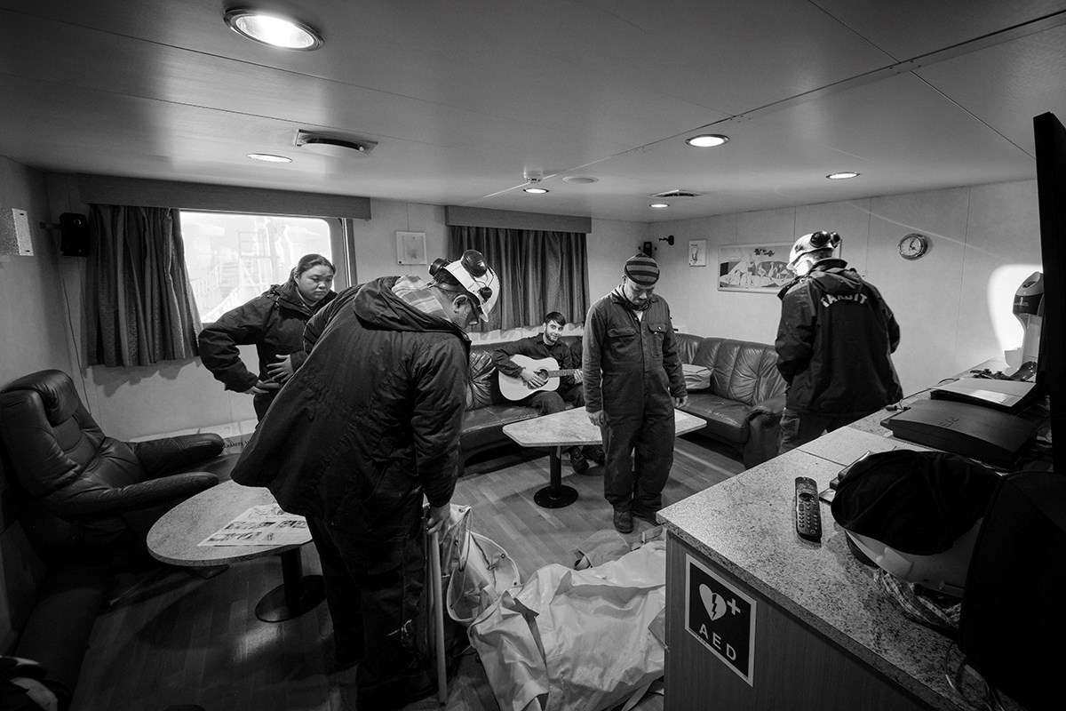 A black‑and‑white photo of four people standing in the ship’s TV room, looking at what appears to be a stretcher or other rescue equipment, while another person sits on the sofa in the background playing a guitar