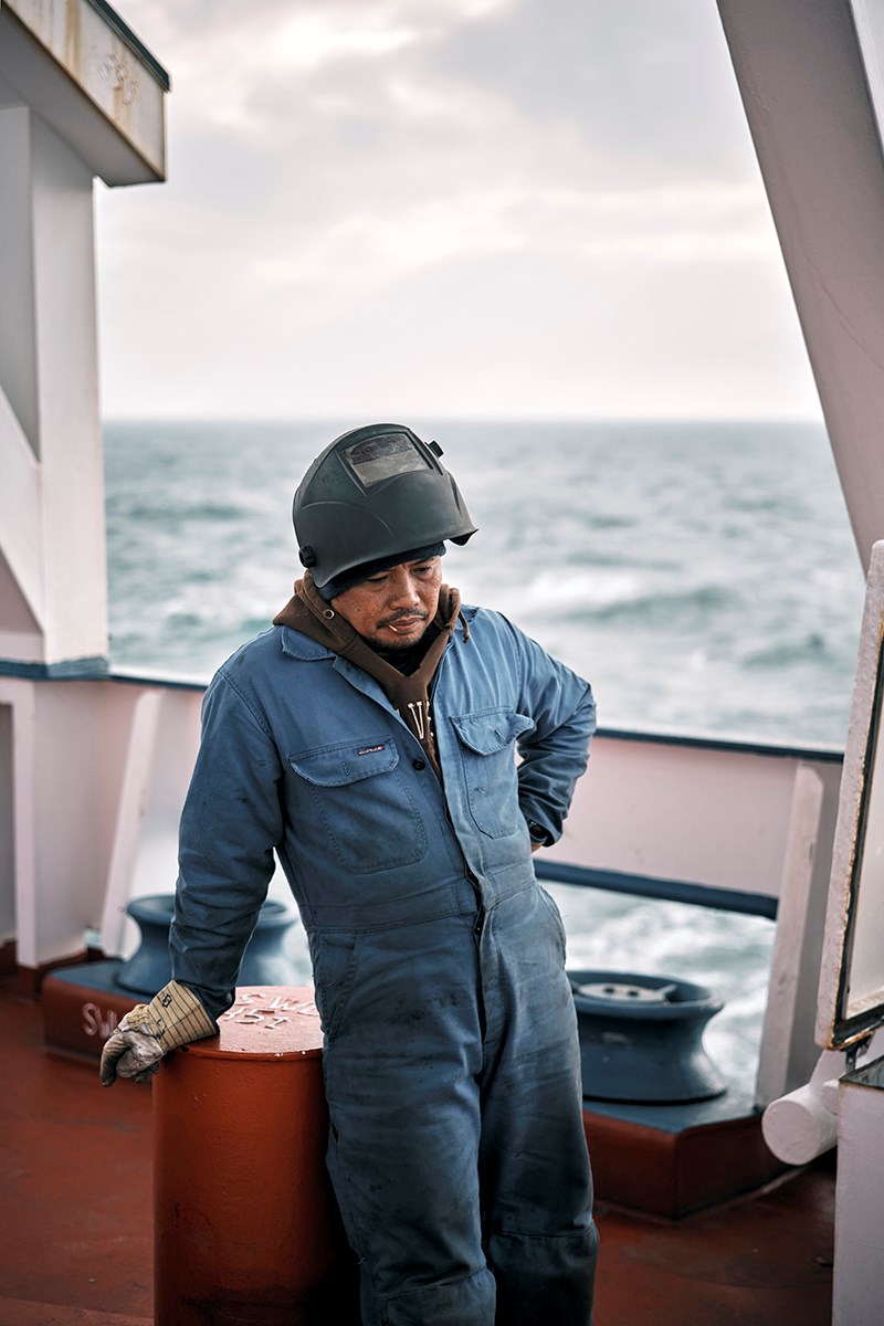 A man wearing a blue coverall and a welding mask pushed up on his head leans against one of the ship’s bollards, a toothpick in the corner of his mouth
