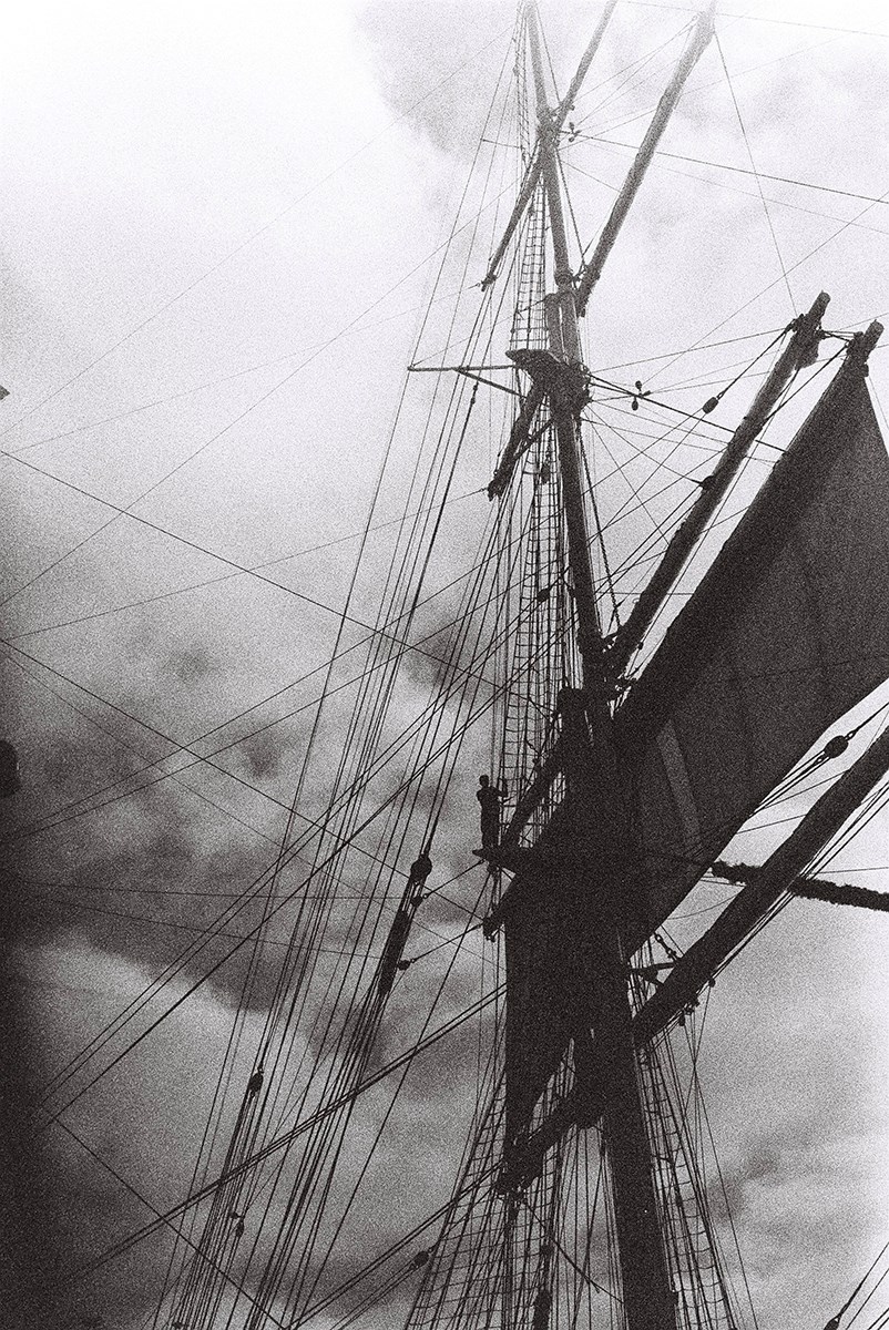 A dramatic black‑and‑white image showing the silhouette of a person high up in the mast of a sailing ship