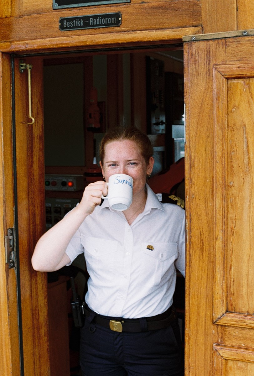 A woman stands in the doorway of the radio room on an older ship. She holds a cup to her mouth, but her eyes reveal a smile