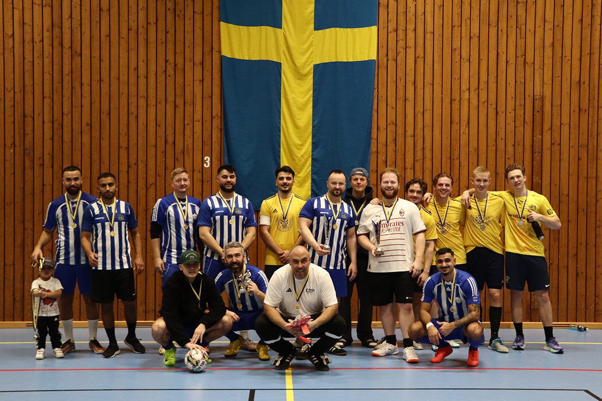 En grupp fotbollsspelare står uppställda mot en träpanelsvägg, framför en stor Sverigeflagga